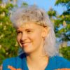 [ALT_TEXT: Una joven con un corte mullet rubio platino rizado, sentada en un parque en un día soleado.]