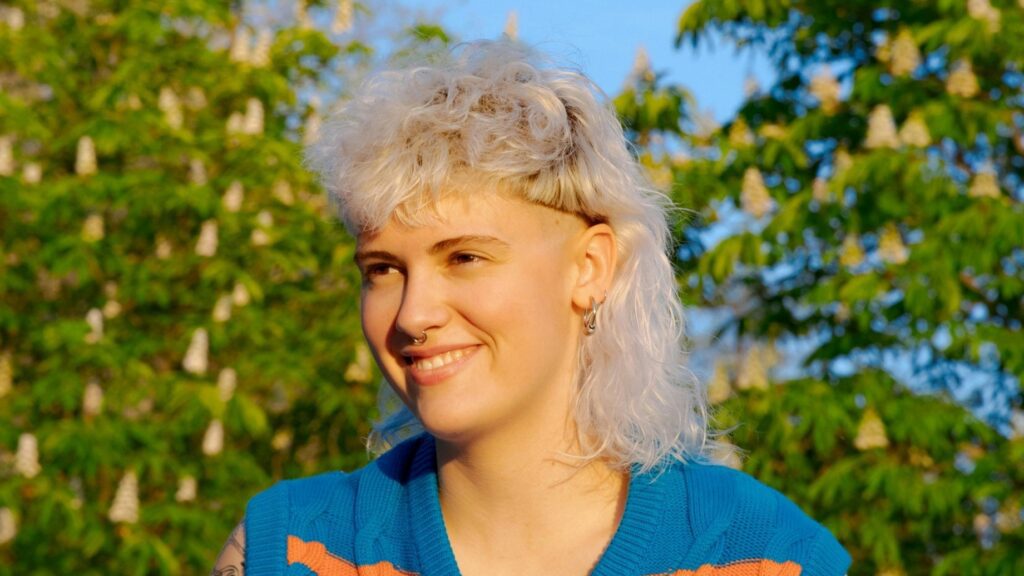 [ALT_TEXT: Una joven con un corte mullet rubio platino rizado, sentada en un parque en un día soleado.]
