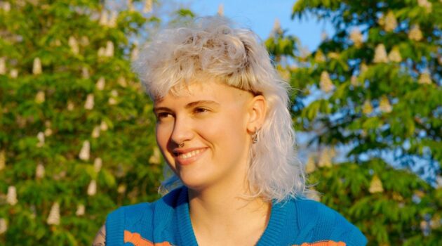 [ALT_TEXT: Una joven con un corte mullet rubio platino rizado, sentada en un parque en un día soleado.]