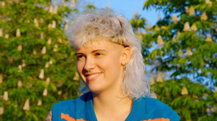 [ALT_TEXT: Una joven con un corte mullet rubio platino rizado, sentada en un parque en un día soleado.]
