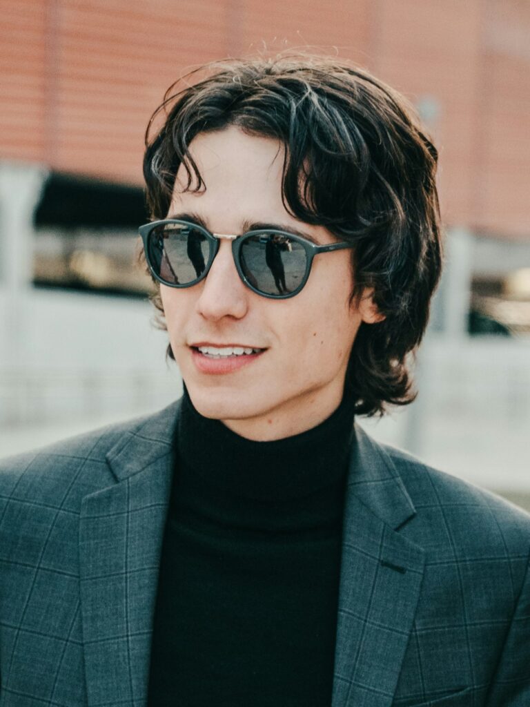 [ALT_TEXT: Un joven con un corte mullet retro, vestido con chaqueta gris y gafas de sol, sonriendo al aire libre.]