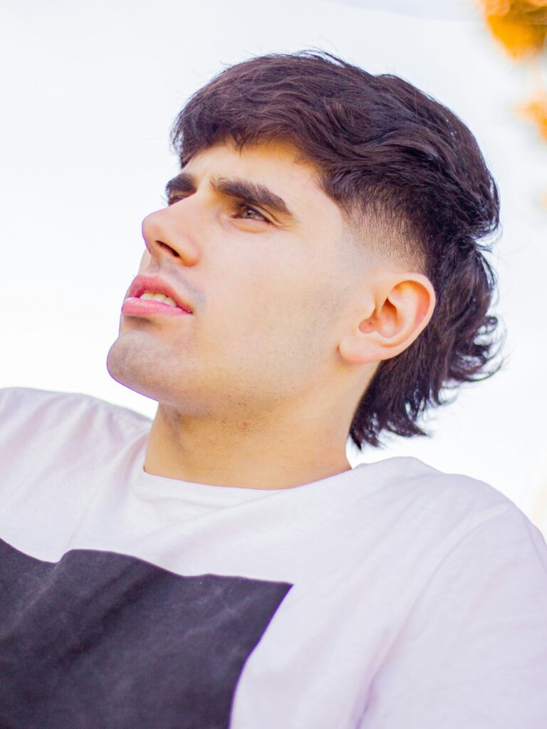 [ALT_TEXT: Un joven con un corte mullet con degradado marcado, vestido con camiseta blanca, sentado junto a una carretera en un día luminoso.]