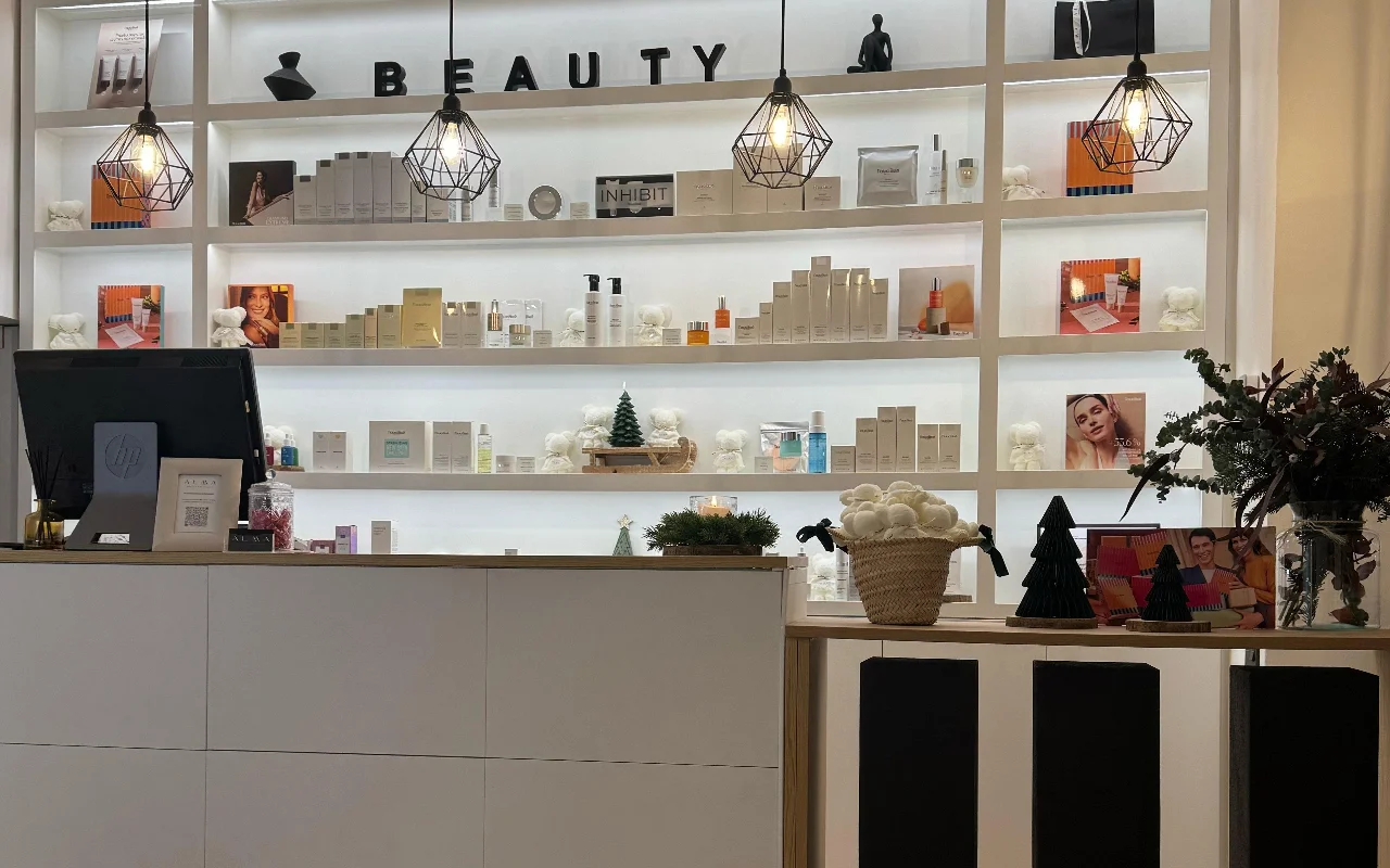 Una recepción luminosa y moderna con una gran estantería blanca retroiluminada llena de productos para el cuidado de la piel. Del techo cuelgan lmparas geométricas de alambre negro, y el mostrador es un mueble blanco minimalista.