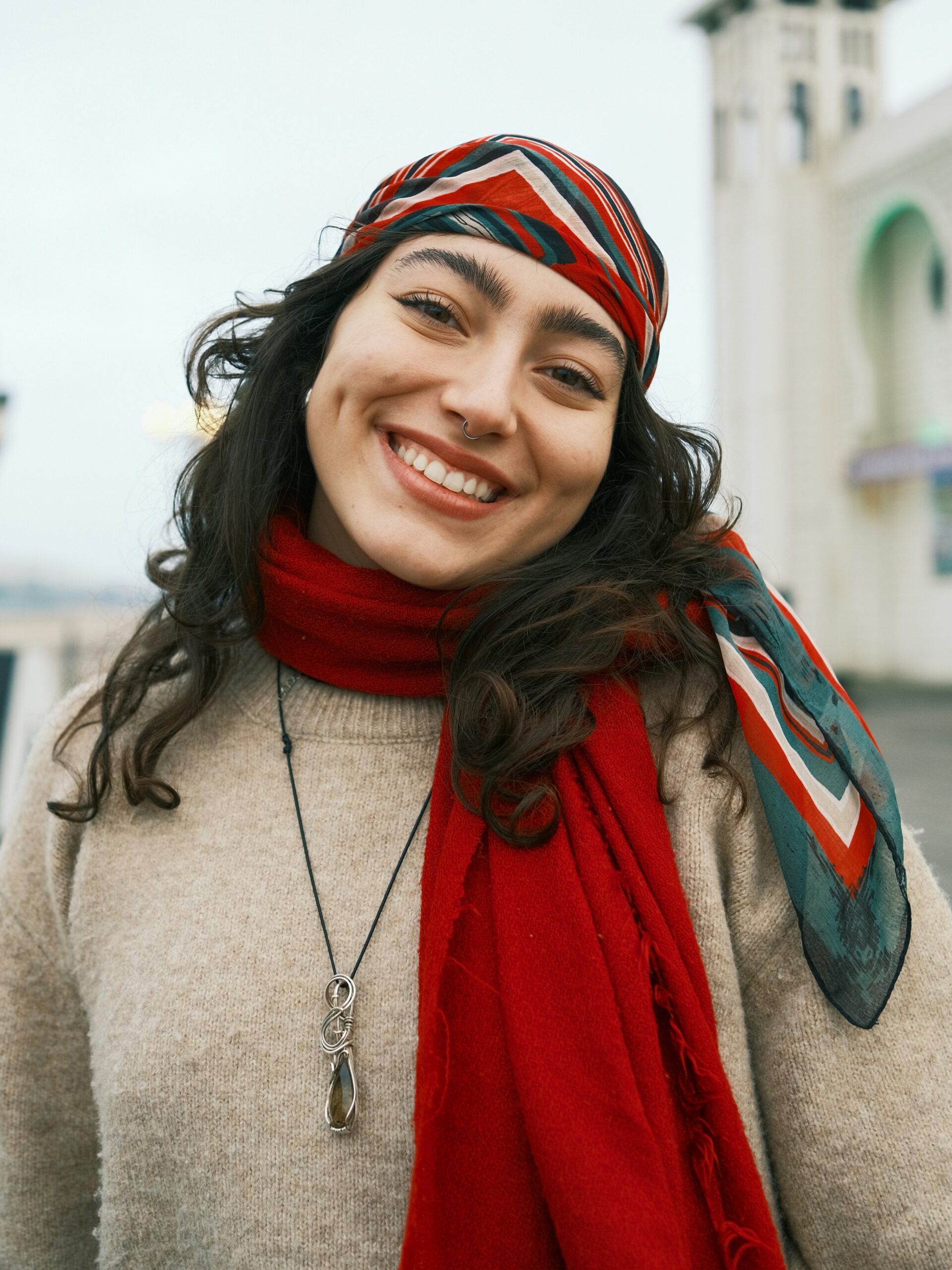 Mujer sonriente de pie en un muelle, con pelo largo oscuro, un pañuelo de rayas rojas, blancas y verdes en la cabeza y un pañuelo rojo al cuello