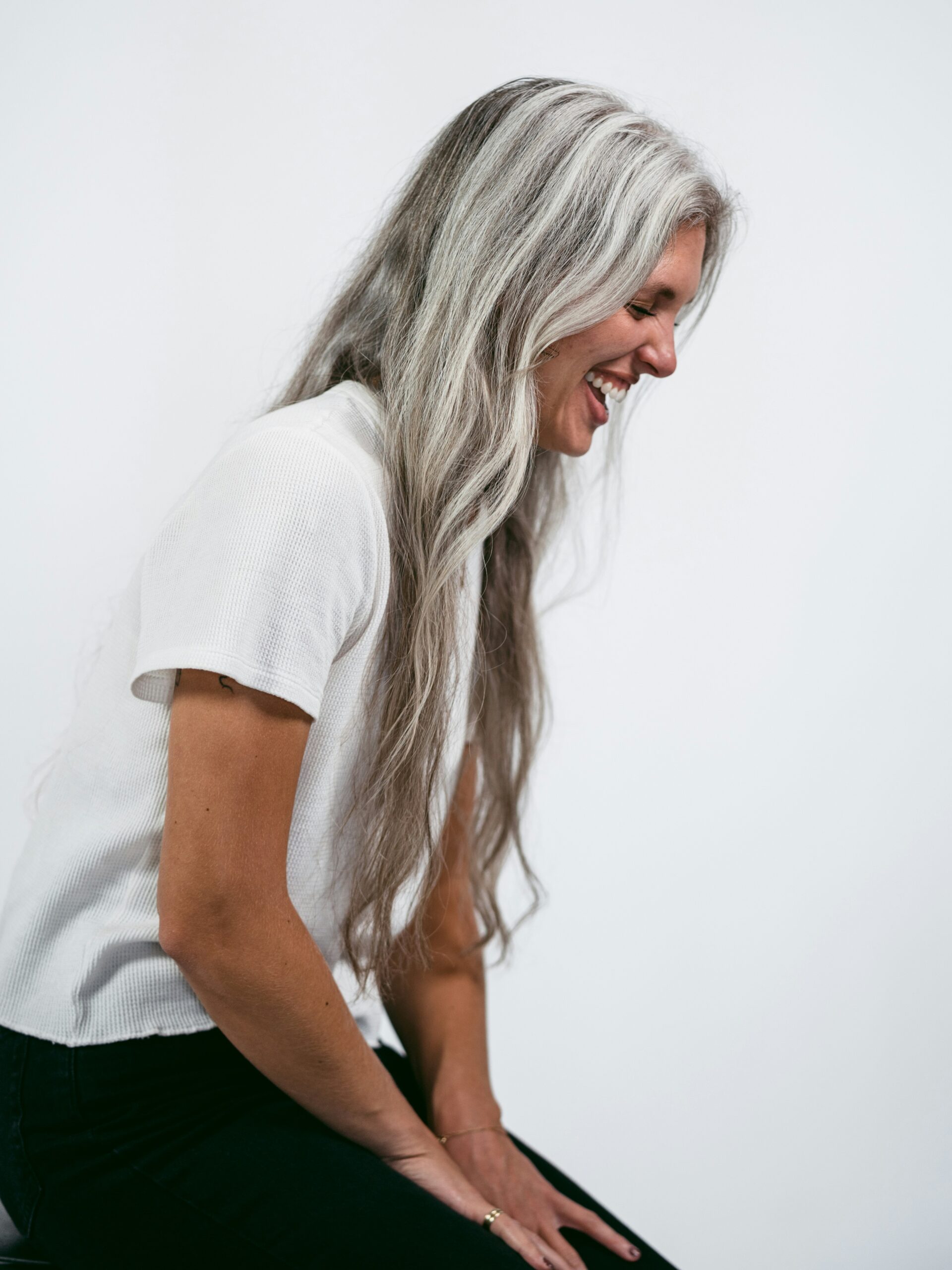 Vista lateral de una mujer sentada en un taburete, riendo, con el pelo gris muy largo