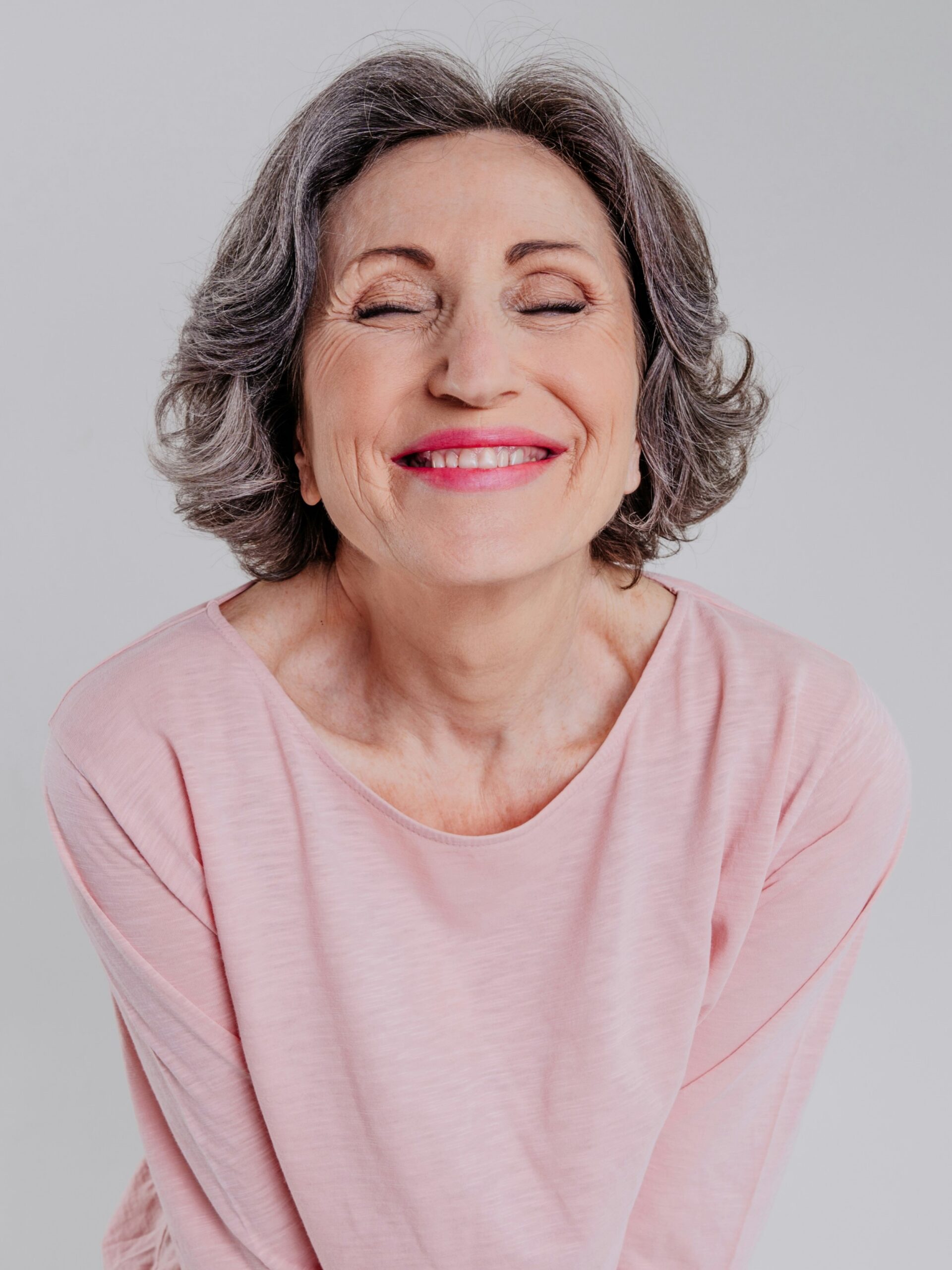 Retrato de una mujer con cabello corto ondulado sal y pimienta, blusa rosa holgada, sonriendo con los ojos cerrados
