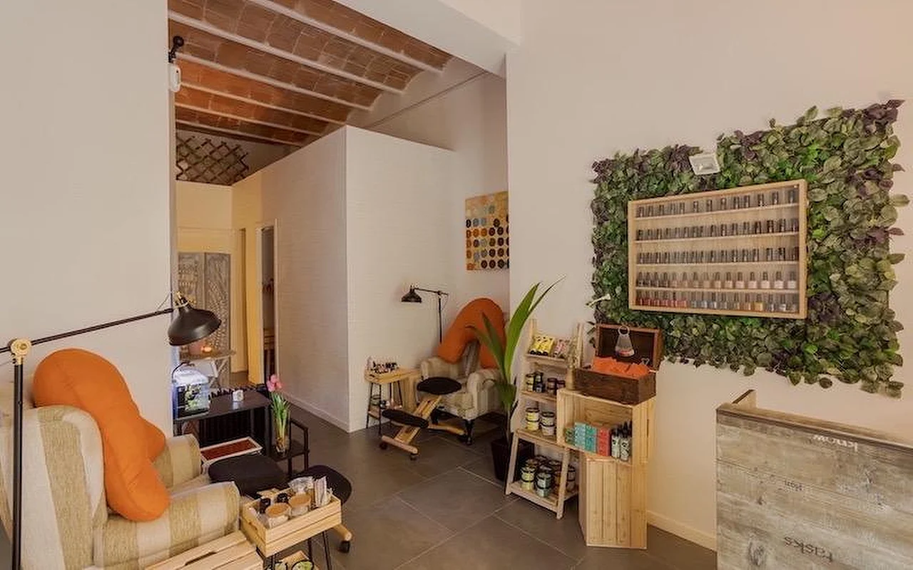 Un salón de uñas acogedor de estilo bohemio, con sillones mullidos y cojines naranjas. La pared muestra una gran instalación vertical de plantas que rodea una vitrina de madera llena de esmaltes de uñas.