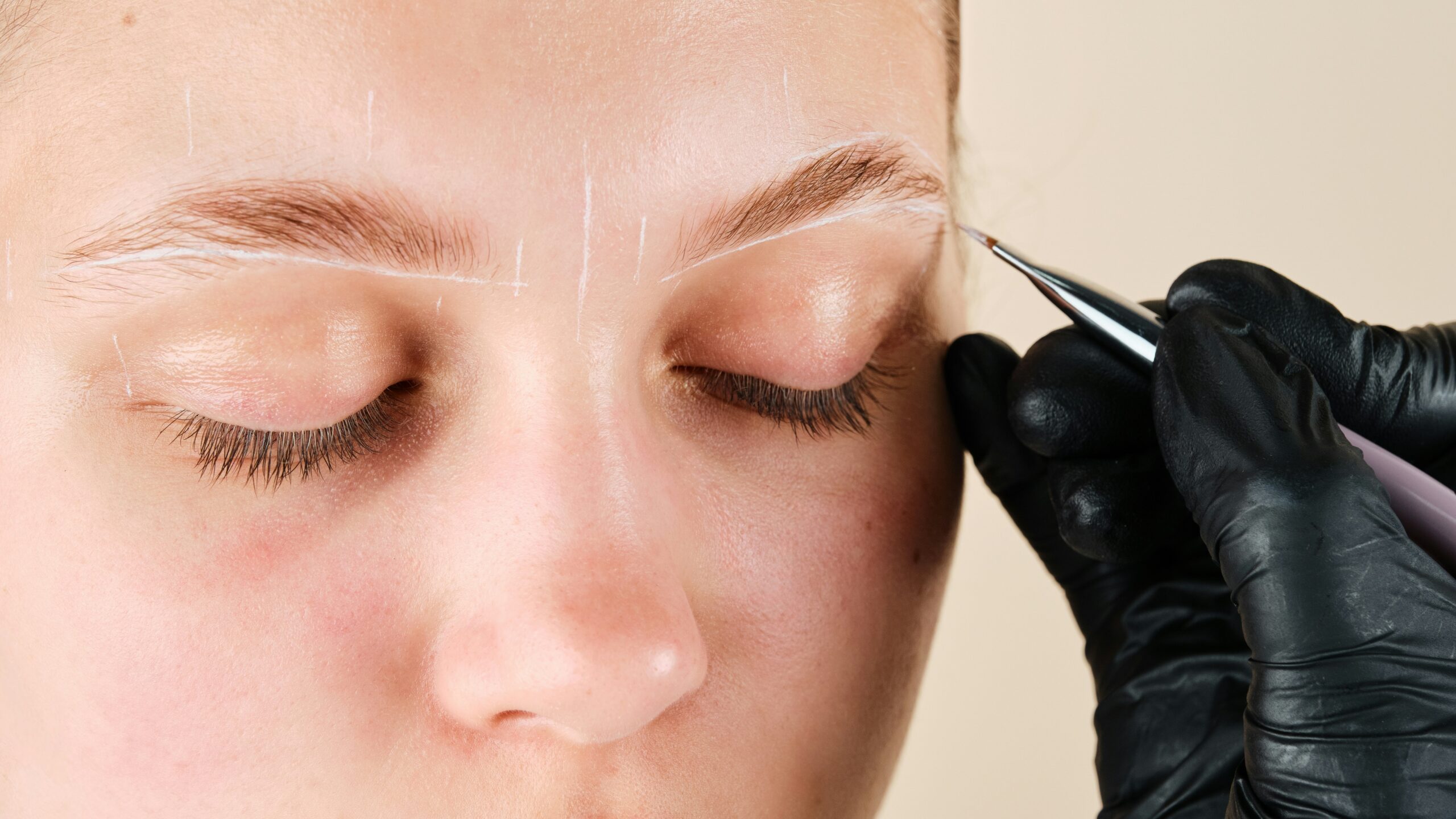 Primer plano del rostro de una mujer mientras una profesional dibuja marcas blancas en la cara para mapear la forma deseada de las cejas
