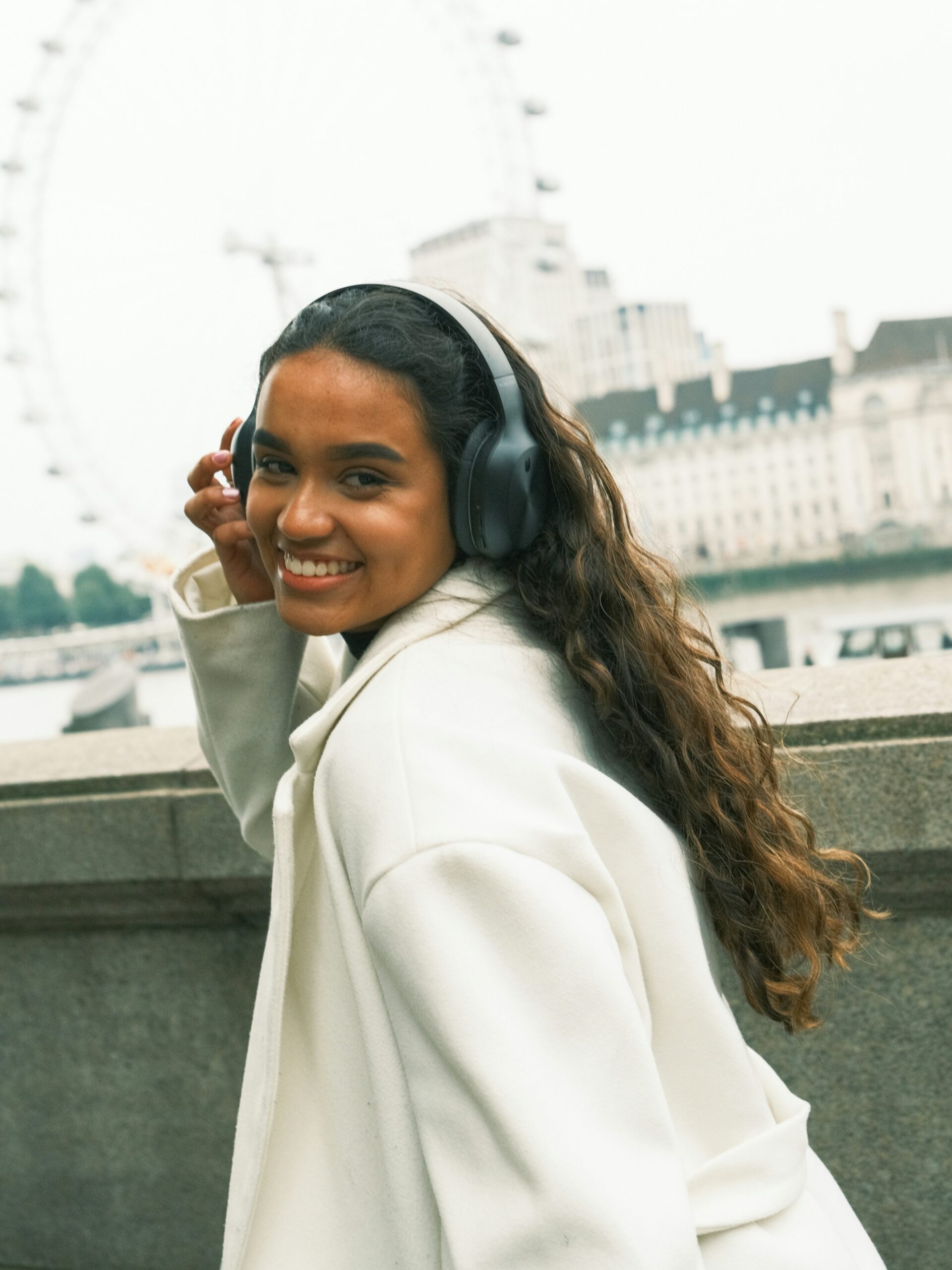 Una mujer sonriente con microblading en las cejas, auriculares negros de diadema y un abrigo blanco paseando junto al río