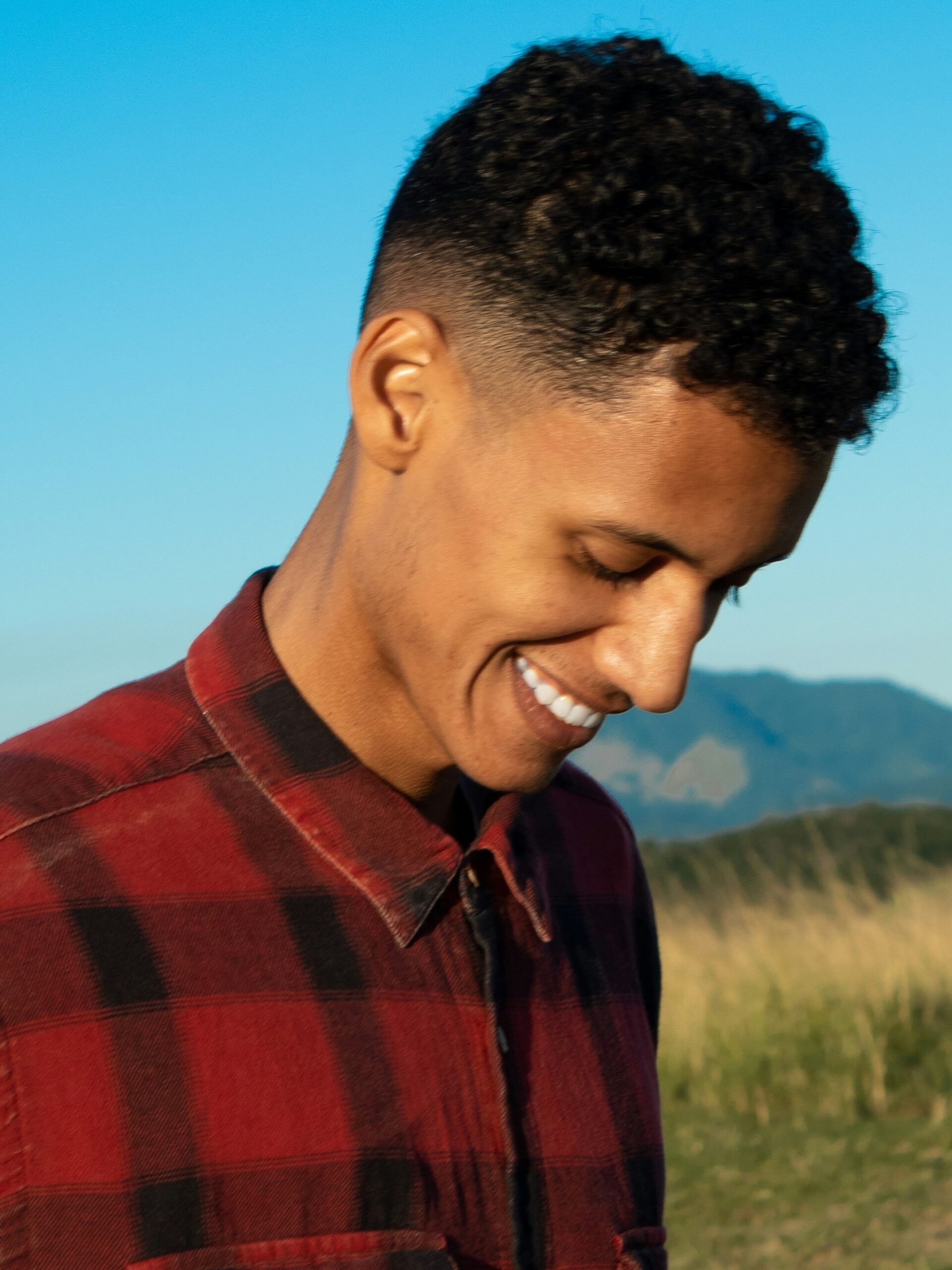 Hombre sonriente al aire libre con montañas y cielo azul de fondo, con cabello rizado y un mid taper fade