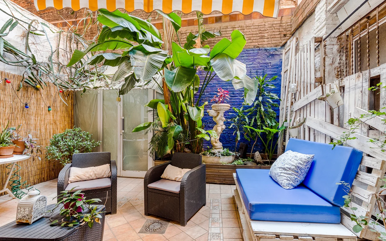 Un patio exterior ecléctico con una llamativa pared azul y abundantes plantas tropicales. Se ven sillones de mimbre oscuro, un banco con cojines azules y un toldo de rayas amarillas y blancas