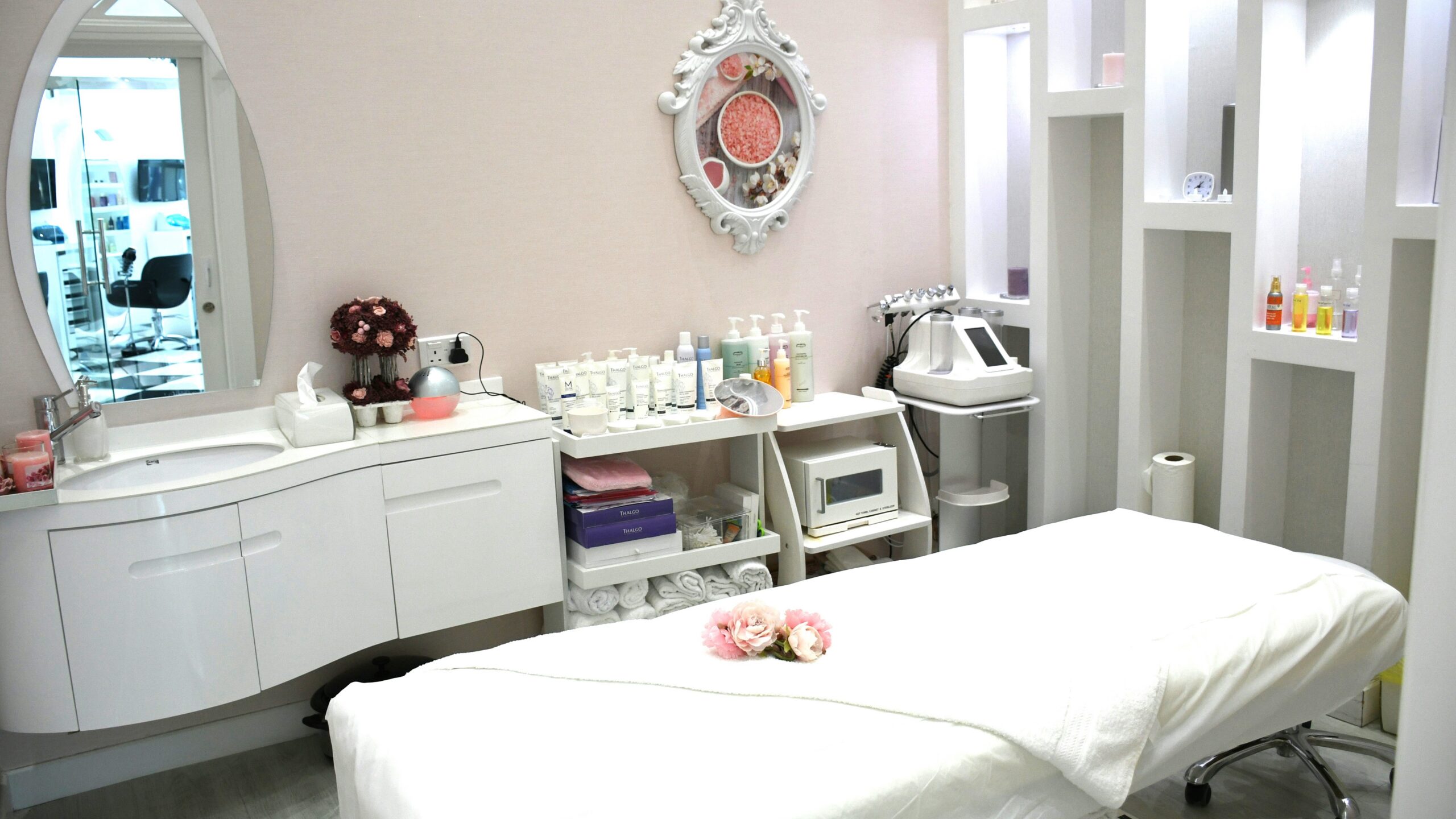 Sala de tratamiento luminosa, limpia y acogedora con una camilla, productos en estanterías, un espejo ornamental y flores