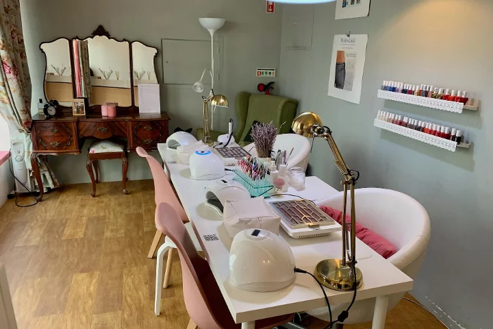 Vista interior de un acogedor salón de belleza especializado en manicuras. En primer plano, una mesa blanca alargada con lámparas de uñas, flexos y herramientas de manicurista, flanqueada por sillas rosas y blancas. Al fondo, un tocador vintage de madera con espejo de tres cuerpos, una butaca verde y baldas blancas en la pared mostrando filas de esmaltes de colores.
