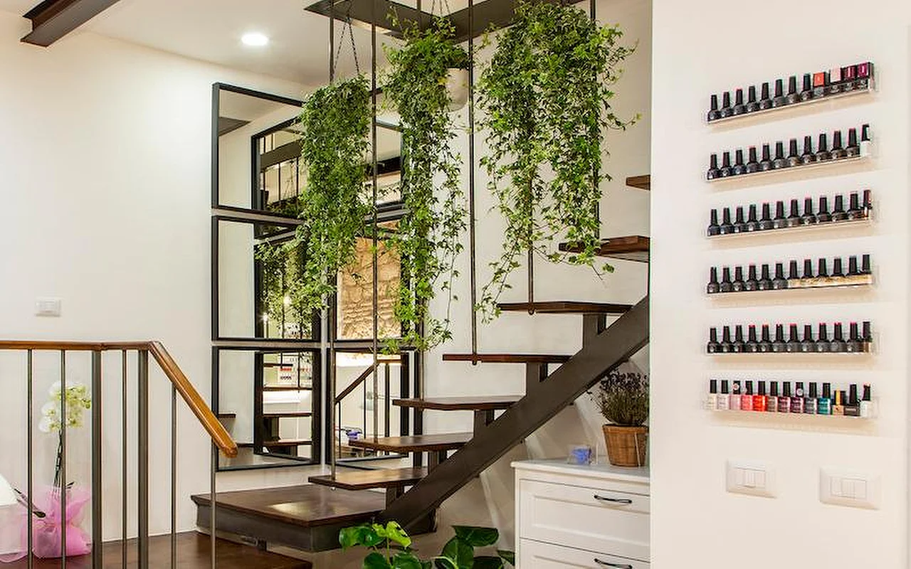Un interior moderno y elegante con una escalera voladiza de madera y metal. Plantas colgantes caen del techo entre los peldaños, y filas de esmaltes de colores se exhiben en estantes transparentes sobre la pared blanca.