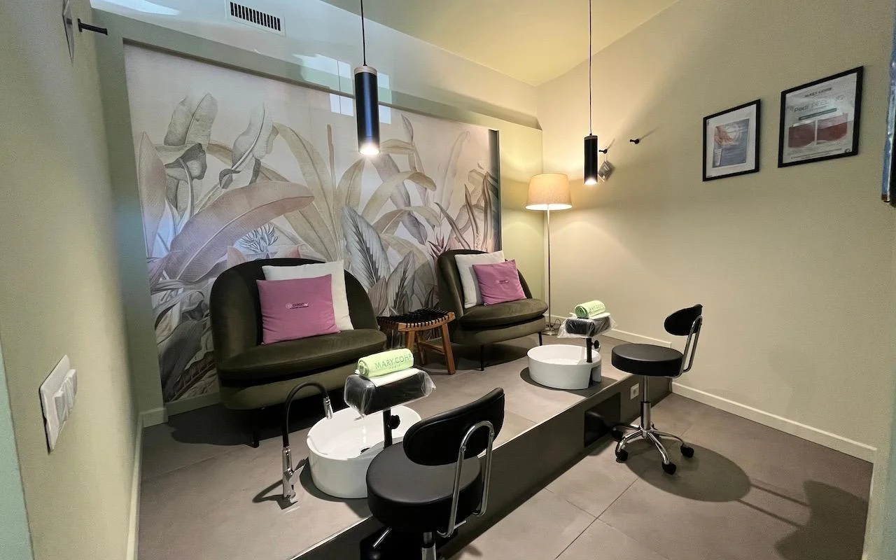 Una estación de pedicura sofisticada con dos sillones de terciopelo verde oscuro y cojines rosa. Baños de pies blancos integrados en una plataforma gris elevada, con un mural de grandes hojas tropicales en la pared del fondo.
