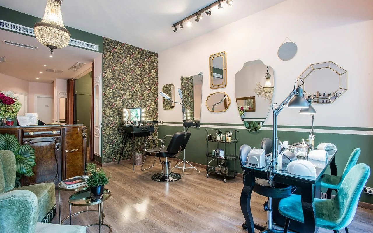 Un salón de estilo vintage-chic con paredes en blanco y verde bosque. Incluye papel pintado floral, un surtido de espejos con marcos dorados de formas irregulares, una estación de maquillaje profesional y asientos de terciopelo verde.