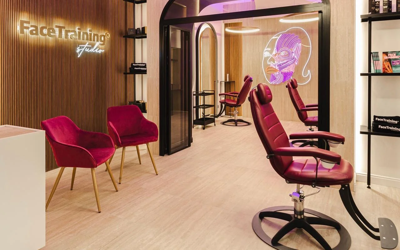 Un estudio minimalista con una pared de lamas de madera y un logotipo retroiluminado. La zona de tratamiento incluye sillones profesionales de piel burdeos y una instalación de neón que muestra un diagrama estilizado de la musculatura facial sobre una mampara de cristal.