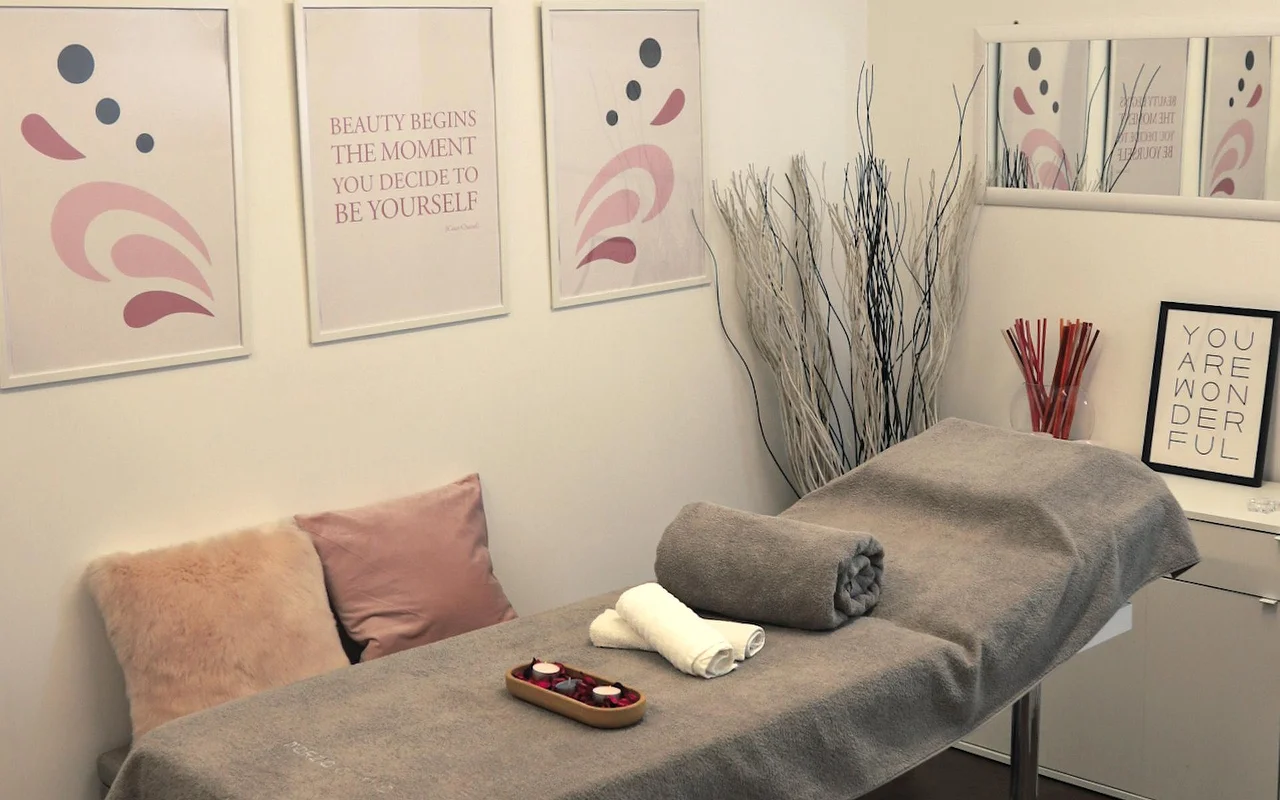 Una sala de tratamiento luminosa con una camilla cubierta con una toalla gris. La pared está decorada con arte abstracto en tonos pastel y frases motivacionales, mientras que ramas blancas decorativas y velas crean una atmósfera de calma.