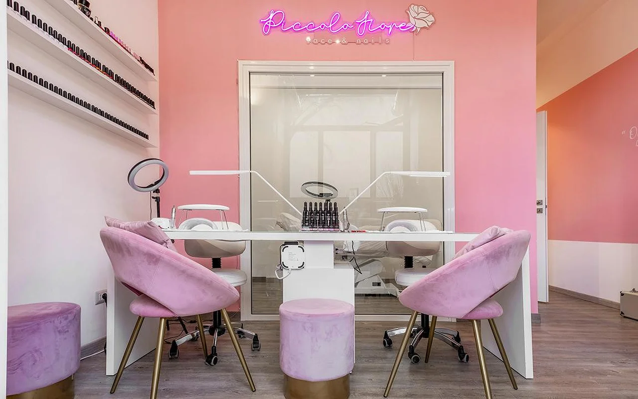 Una estación de manicura con paredes rosa intenso y un neón de color fucsia. Sillas de terciopelo lila con patas doradas junto a un escritorio blanco minimalista equipado con lámparas LED profesionales para uñas.