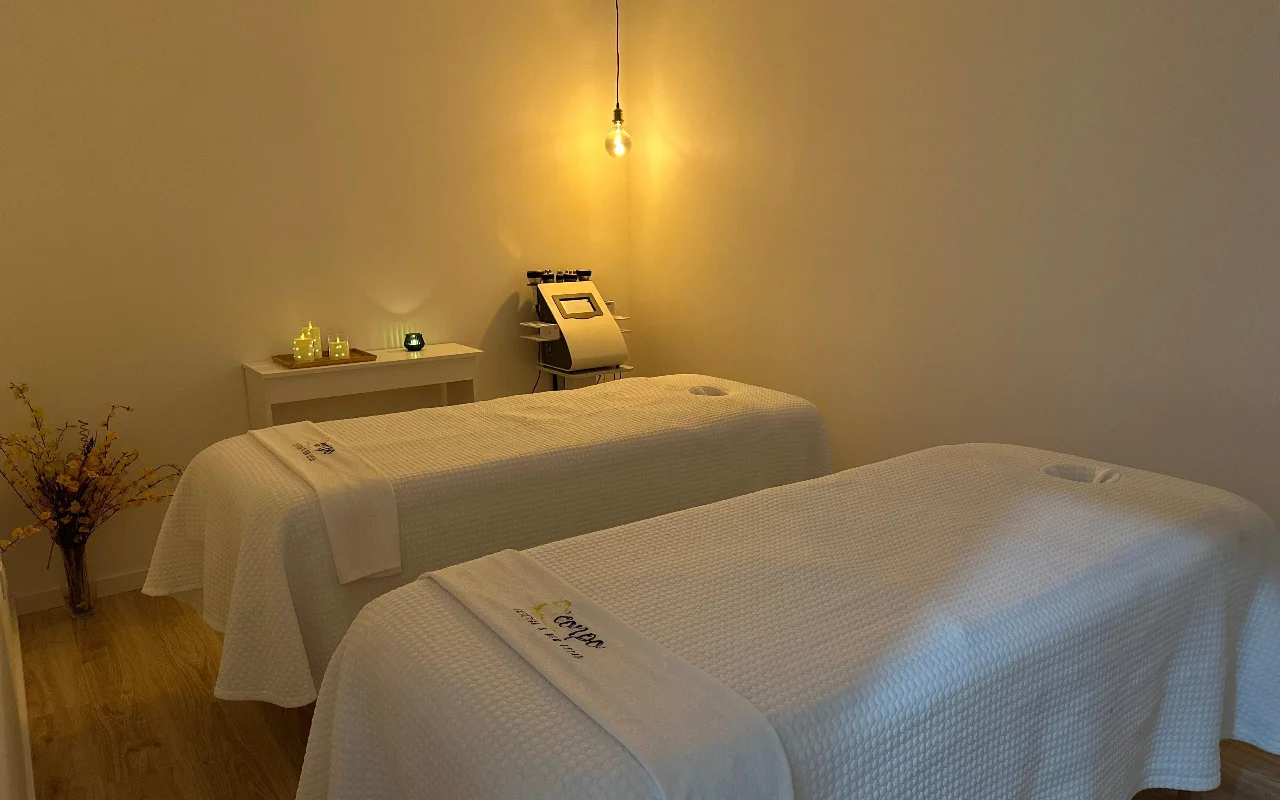 Una tranquila sala de masaje o tratamiento con dos camillas colocadas una junto a otra, vestidas con toallas y sábanas blancas impecables. La sala está iluminada por una única bombilla colgante de luz cálida y pequeñas velas sobre una mesa auxiliar, creando una atmósfera serena. Al fondo, entre las camillas, una máquina cosmética especializada.