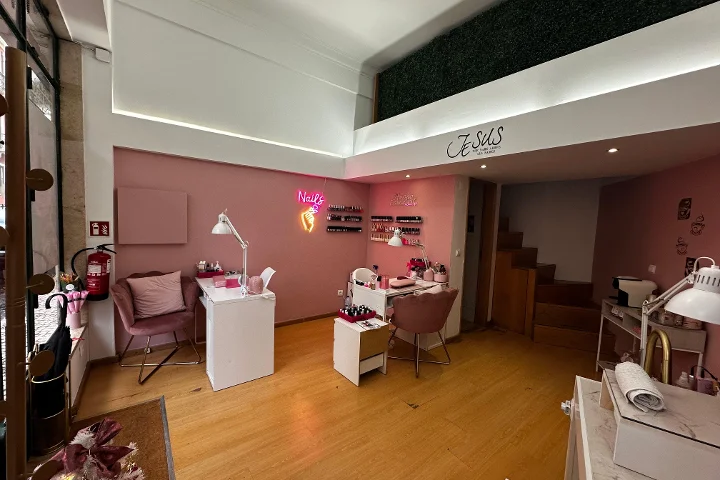 Un estudio de uñas con encanto, paredes en rosa claro y suelo de madera cálida. La sala tiene varias mesas blancas de manicura con sillas de terciopelo en rosa palo. Un cartel de neón rosa con forma de mano sosteniendo un bote de esmalte y la palabra 'Nails' brilla en la pared, y filas de esmaltes se exponen en pequeñas baldas. En la parte superior se ve un altillo con una pared de seto artificial verde.