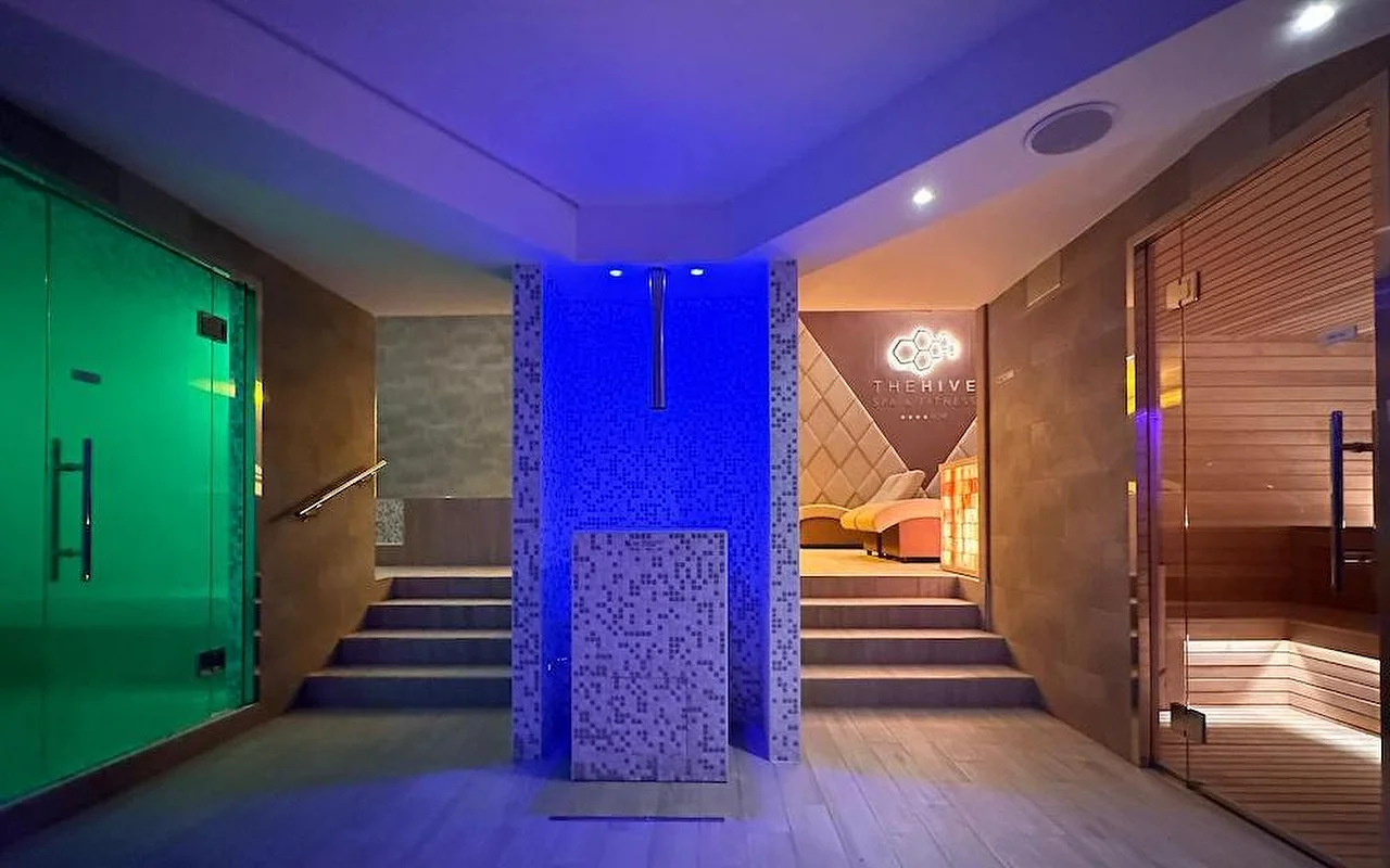 La zona termal de un spa de lujo con una iluminación atmosférica en tonos azules y verdes. El espacio incluye una sauna finlandesa de madera, una entrada al baño de vapor y un mosaico central con escalones que conducen a zonas de descanso.