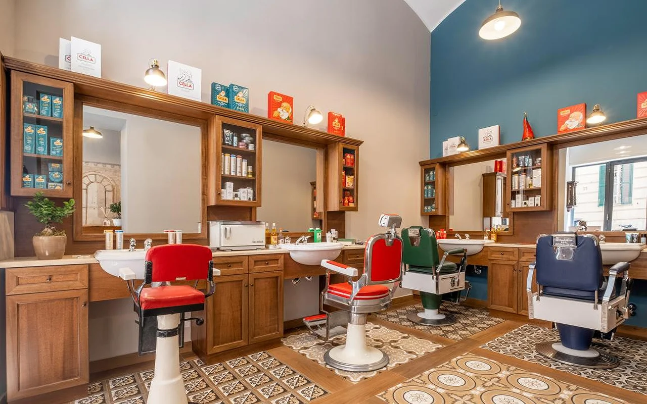 Una barbería tradicional de alta gama con muebles de madera noble, baldosas con motivos vintage en el suelo y sillones de barbero en rojo, verde y azul marino. Productos de aseo clásicos se exhiben en las estanterías.