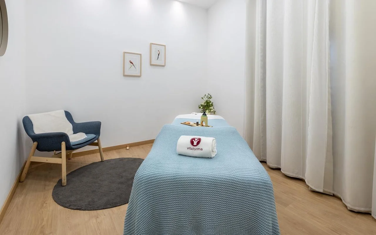 Una sala de spa limpia y minimalista, con paredes blancas y suelo de madera clara. Una camilla de tratamiento está cuidadosamente preparada con una manta de punto azul claro y una toalla blanca enrollada con el logo de 'Vitaforma'. En un rincón, una butaca azul elegante sobre una pequeña alfombra redonda gris, y largas cortinas blancas junto a la ventana.