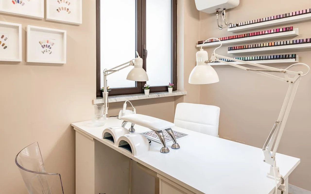 Una estación de manicura dedicada en AG The Beauty Room's, con un escritorio blanco impoluto, dos lámparas UV, luces de trabajo regulables y estanterías que muestran hileras de esmaltes de colores. En las paredes de tonos cálidos cuelgan diseños de nail art enmarcados.