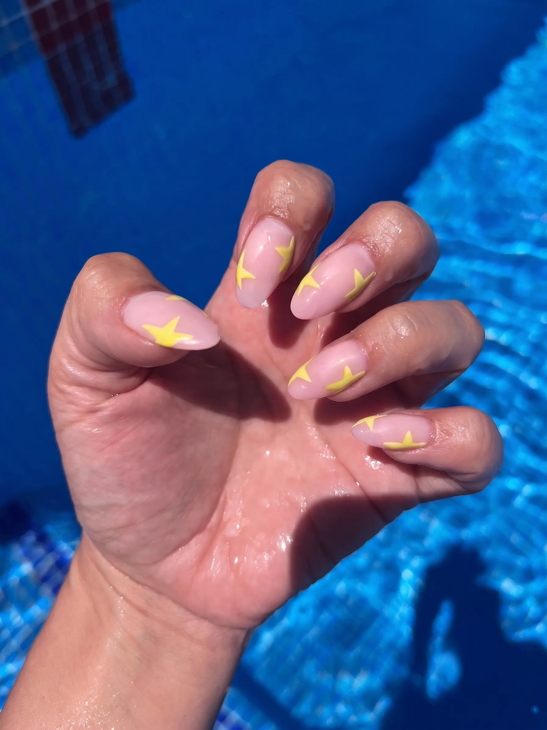 Uñas almendradas en nude transparente con estrellas en amarillo mantequilla, fotografiadas junto a la piscina.