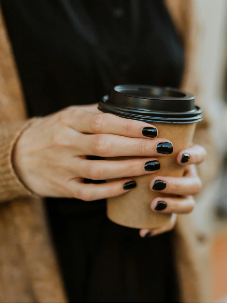Primer plano de una mujer con una manicura de gel negra y brillante sosteniendo un vaso de café para llevar