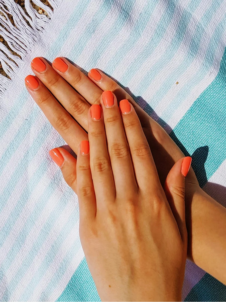 Vista cenital de dos manos con una manicura de gel naranja y brillante apoyadas sobre una toalla de playa de rayas azules y blancas