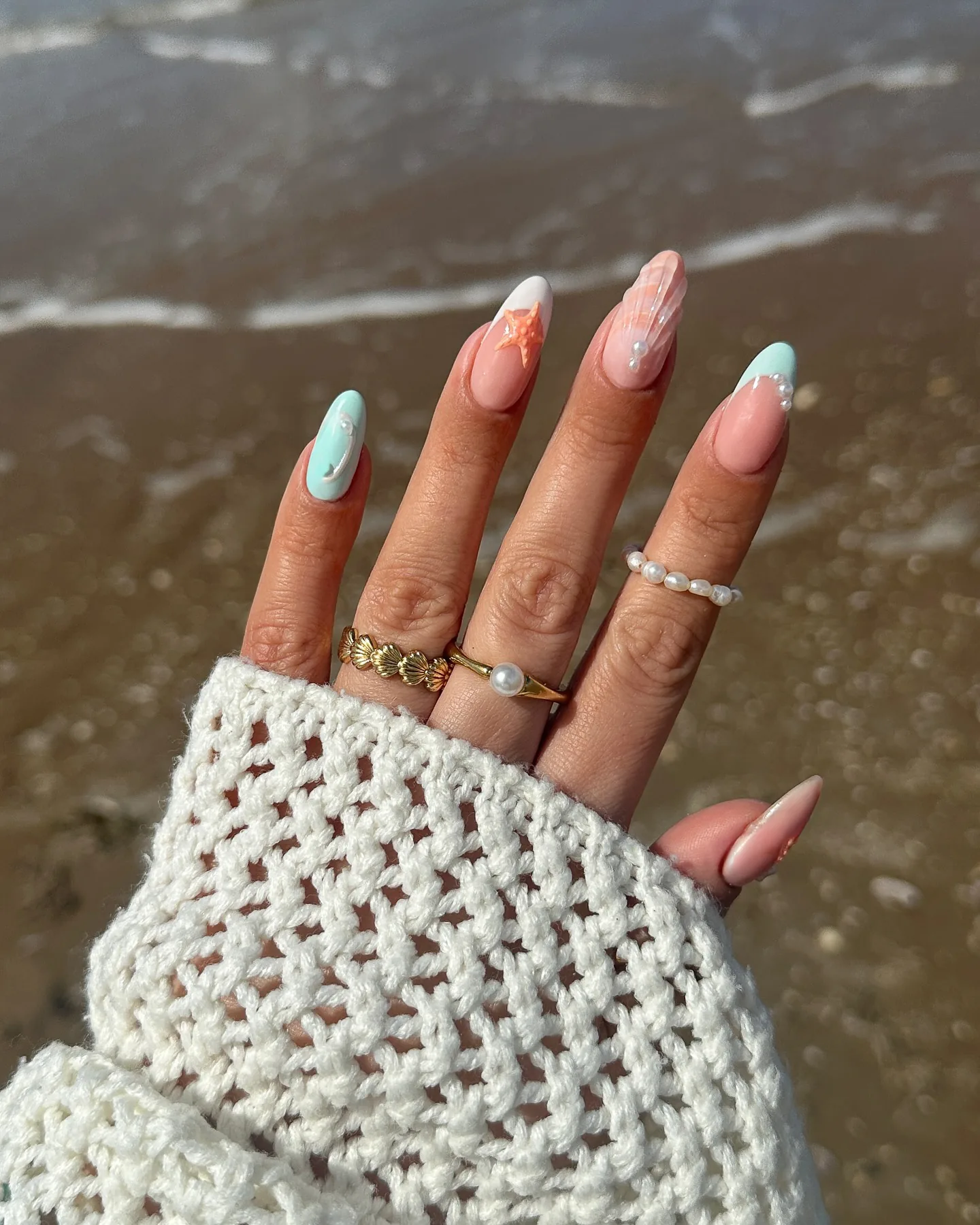 Uñas almendradas con nail art sirena en tonos pastel –una estrella de mar, acentos nacarados y un brillo iridiscente– sobre arena mojada.