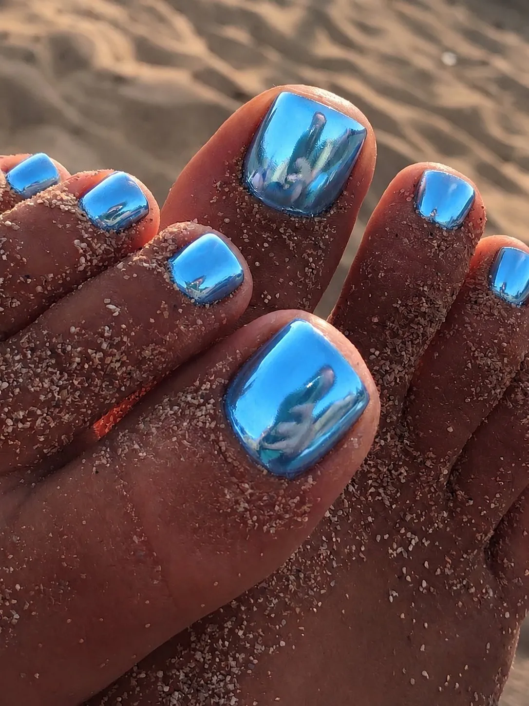 Pedicura cromada azul océano metalizada en unos pies sobre la arena, en la playa