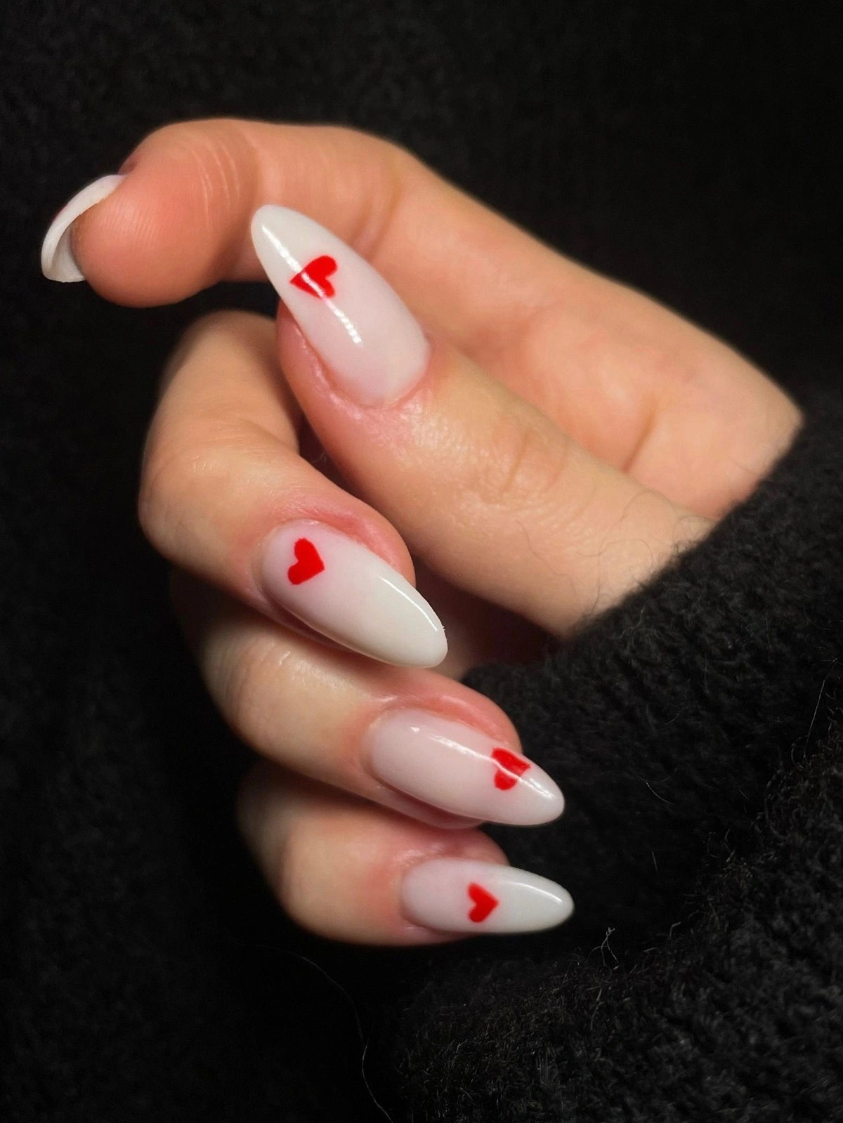 Primer plano de la mano de una mujer con uñas acrílicas almendradas blancas decoradas con pequeños corazones rojos
