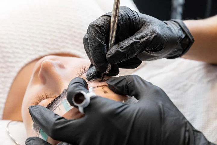 Primer plano de un tratamiento de cejas en marcha: una técnica con guantes negros mide y marca la ceja de una clienta reclinada con una regla específica y un lápiz, los ojos cerrados.