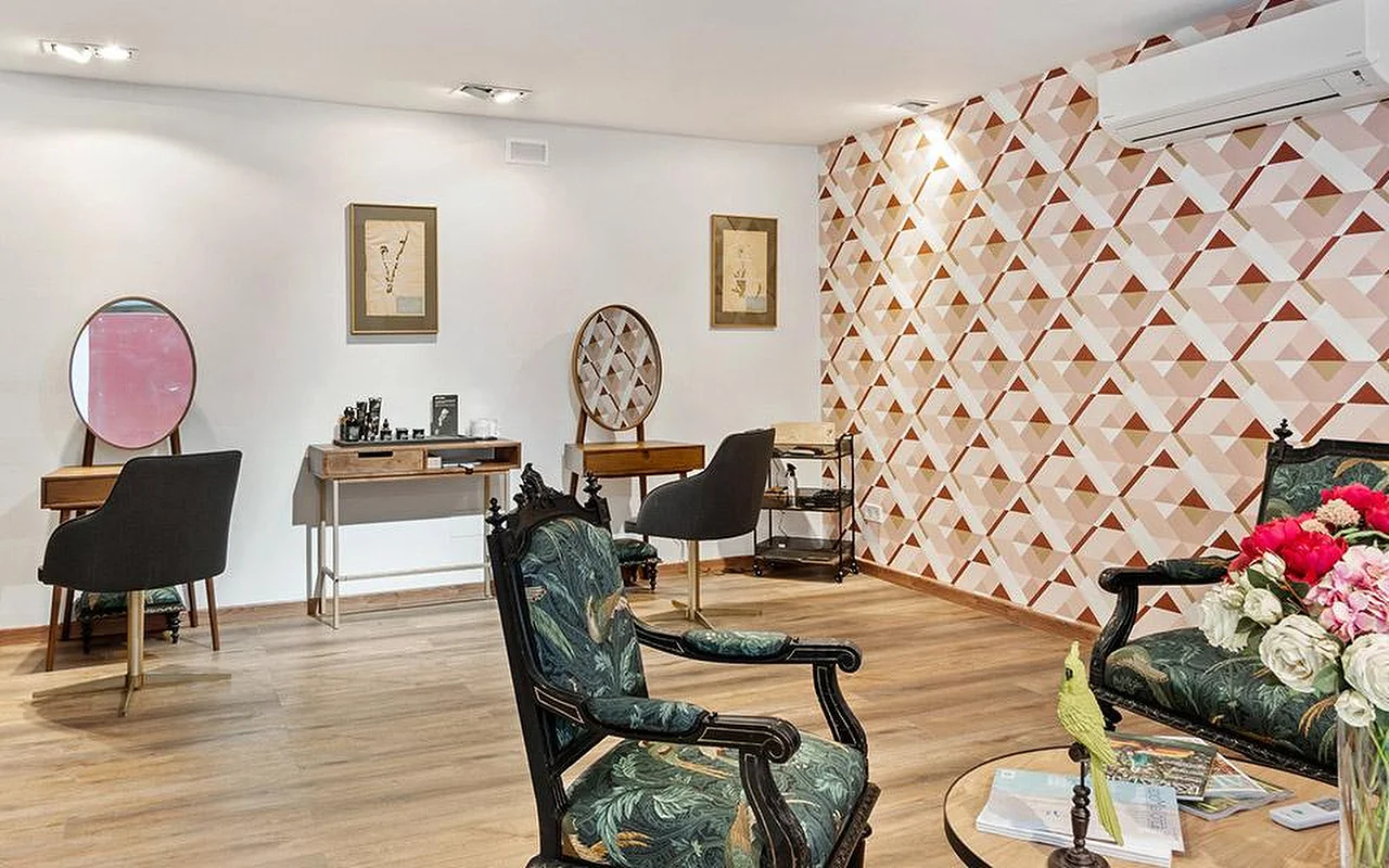 Una elegante sala de salón con una pared decorativa geométrica en rosa y terracota, un par de sillas talladas y ornamentadas tapizadas con tela botánica verde, tocadores de madera con espejos redondos y un jarrón con flores rosas y rojas en primer plano.
