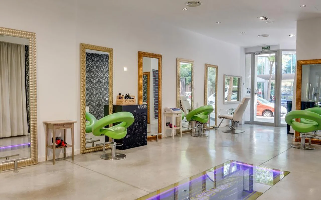 Una peluquería luminosa y espaciosa con suelos de hormigón pulido, altos espejos con marco dorado a lo largo de una pared blanca, sillones de peinado moldeados en verde lima y una zona de suelo de cristal iluminada con luz violeta, con la calle visible a través de las puertas acristaladas del fondo.