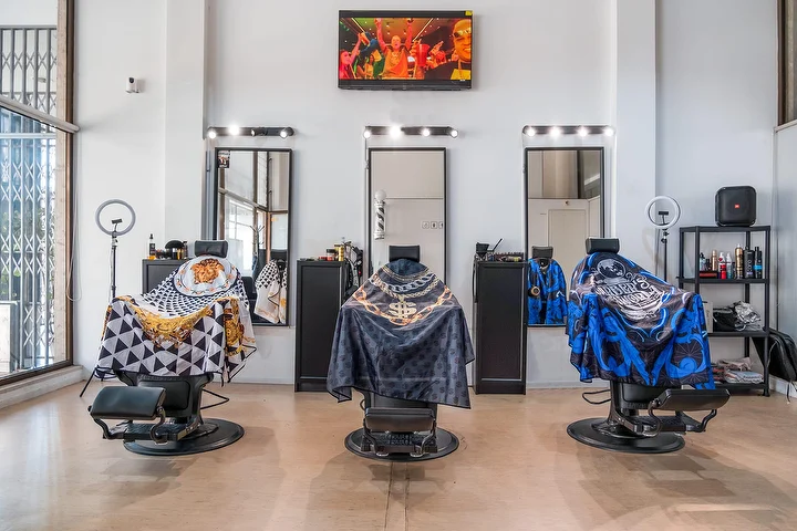 Una barbería luminosa de techos altos con tres sillones negros cubiertos con capas estampadas, cada uno frente a un espejo enmarcado por bombillas, con aros de luz, una pantalla en la pared y ventanales altos.