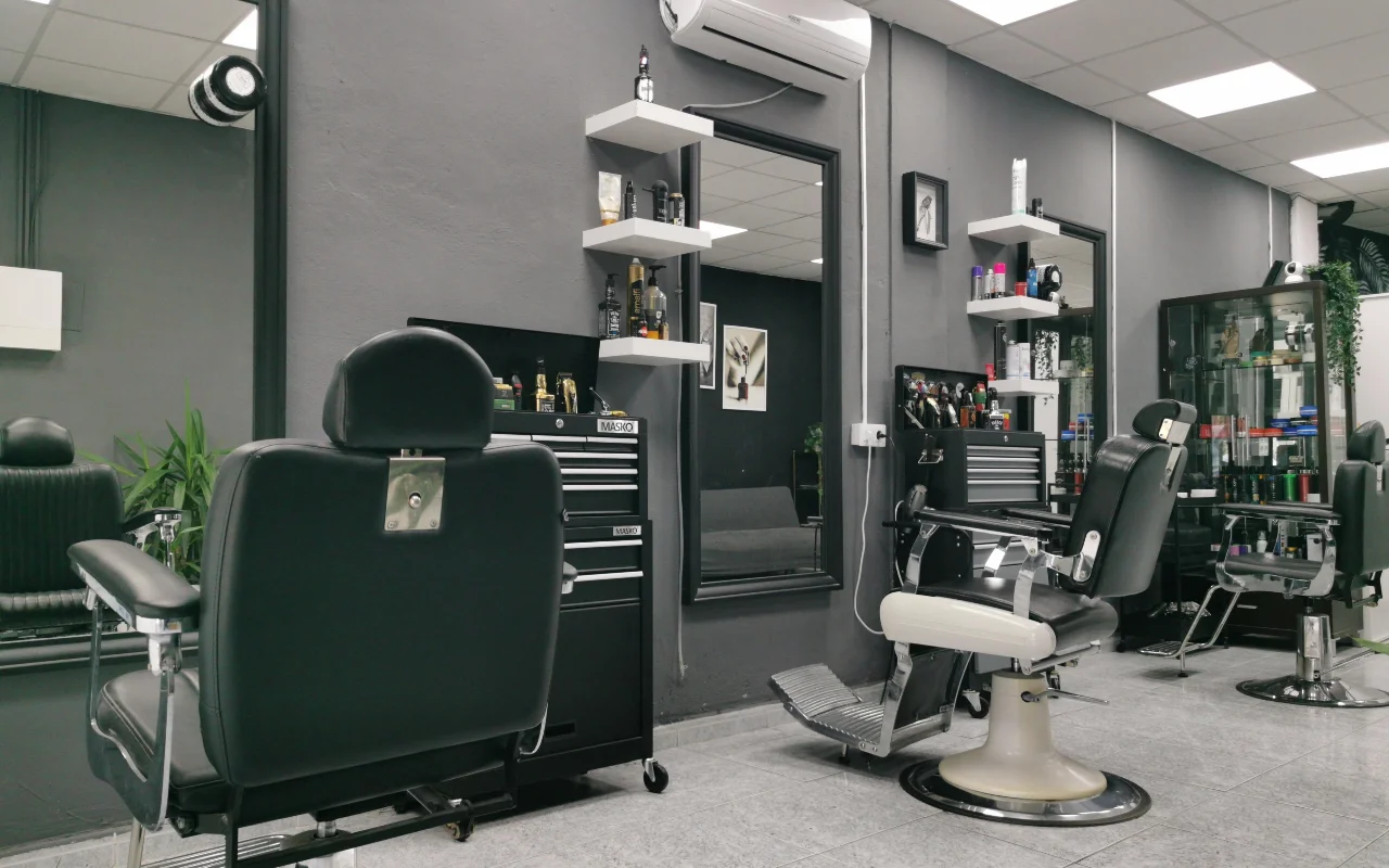 Una barbería tradicional con paredes en gris carbón, clásicos sillones de barbero de cuero negro con bases y reposapiés cromados, baldas blancas flotantes con productos de aseo y grandes espejos sobre suelo de baldosas.