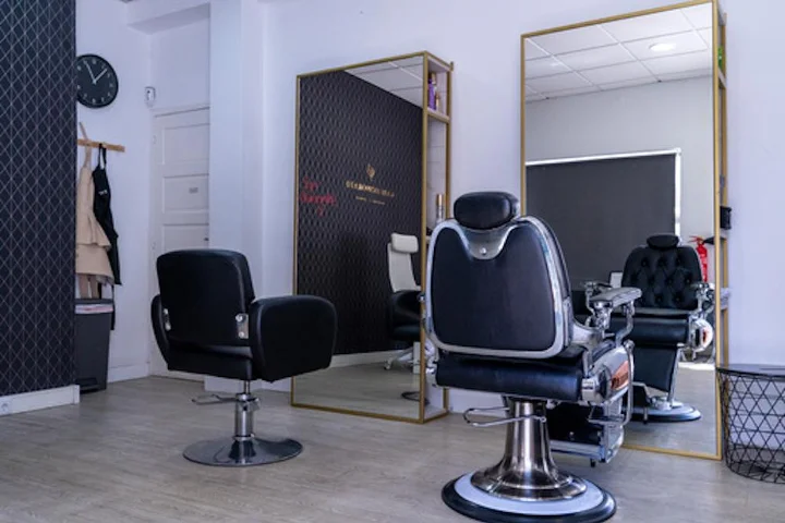 Un interior de salón sobrio con un sillón de peluquería negro y un sillón de barbero de cromo y cuero sobre suelo de madera clara, un gran espejo con marco dorado, un reloj de pared y batas colgadas junto a una pared oscura de acento.