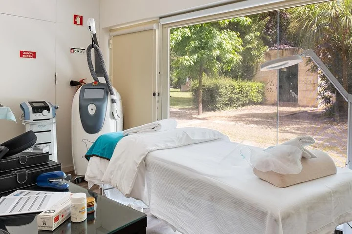Una cabina de tratamientos con una camilla cubierta de blanco junto a un ventanal de suelo a techo que da a un jardín verde, un aparato de tratamientos corporales sobre soporte, una lámpara lupa y una mesa auxiliar de cristal con productos.
