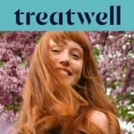 Primer plano de una mujer joven pelirroja y sonriente con flequillo, con el cabello moviéndose al viento frente a un fondo de flores de lila rosas. En la parte superior, una franja azul claro muestra el logotipo de "Treatwell" en azul oscuro.