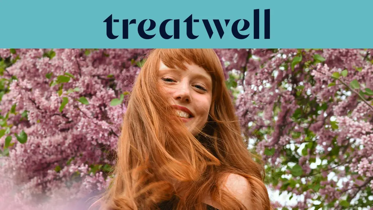 Primer plano de una mujer joven pelirroja y sonriente con flequillo, con el cabello moviéndose al viento frente a un fondo de flores de lila rosas. En la parte superior, una franja azul claro muestra el logotipo de 'Treatwell' en azul oscuro.