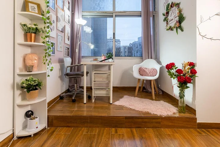 Un pequeño estudio de estética sobre una tarima de madera, con un escritorio blanco y un flexo de pinza, una silla de oficina y una butaca, estanterías de esquina con plantas, un jarrón de rosas rojas y una ventana a la ciudad.