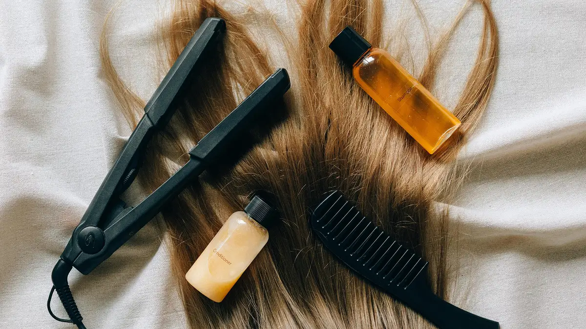 Bodegón de productos para el cuidado del cabello colocados sobre una larga melena lisa, con champú, acondicionador, plancha y un peine de púas anchas