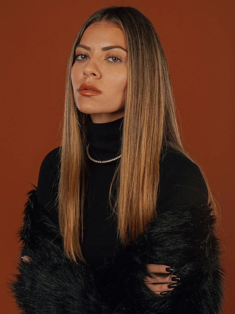 Retrato de estudio de una mujer con una larga melena castaña media, lisa y brillante, con un top negro, una estola de pelo negra y pendientes de aro y collar con brillos