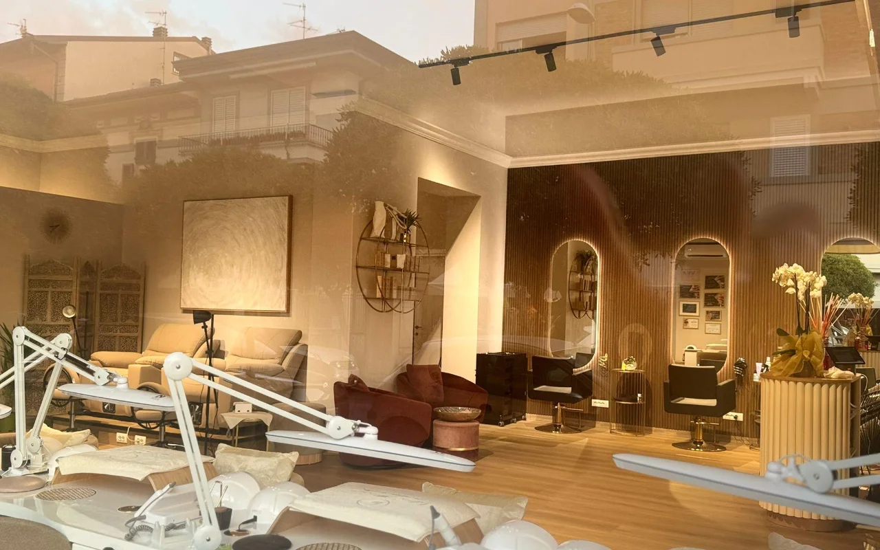 El interior del salón de Lilian Almanza visto a través del escaparate a la hora dorada. Las estaciones de manicura con lámparas regulables se alinean en el lado izquierdo, mientras que el derecho luce paneles de madera cálida, espejos arqueados con marcos retroiluminados y sillones de terciopelo.