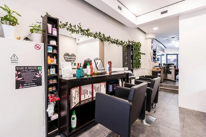Una peluquería estrecha y alargada con sillones negros junto a una pared de espejos, una estantería oscura repleta de productos capilares, una guirnalda de hojas que cae sobre el espejo y un letrero enmarcado de Made In Brazil en una columna blanca.