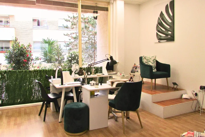 Una zona de manicura luminosa con dos mesas blancas para uñas y sus lámparas, butacas de terciopelo verde y un taburete a juego, un lienzo con hoja de monstera en la pared y grandes ventanales a una terraza con plantas.